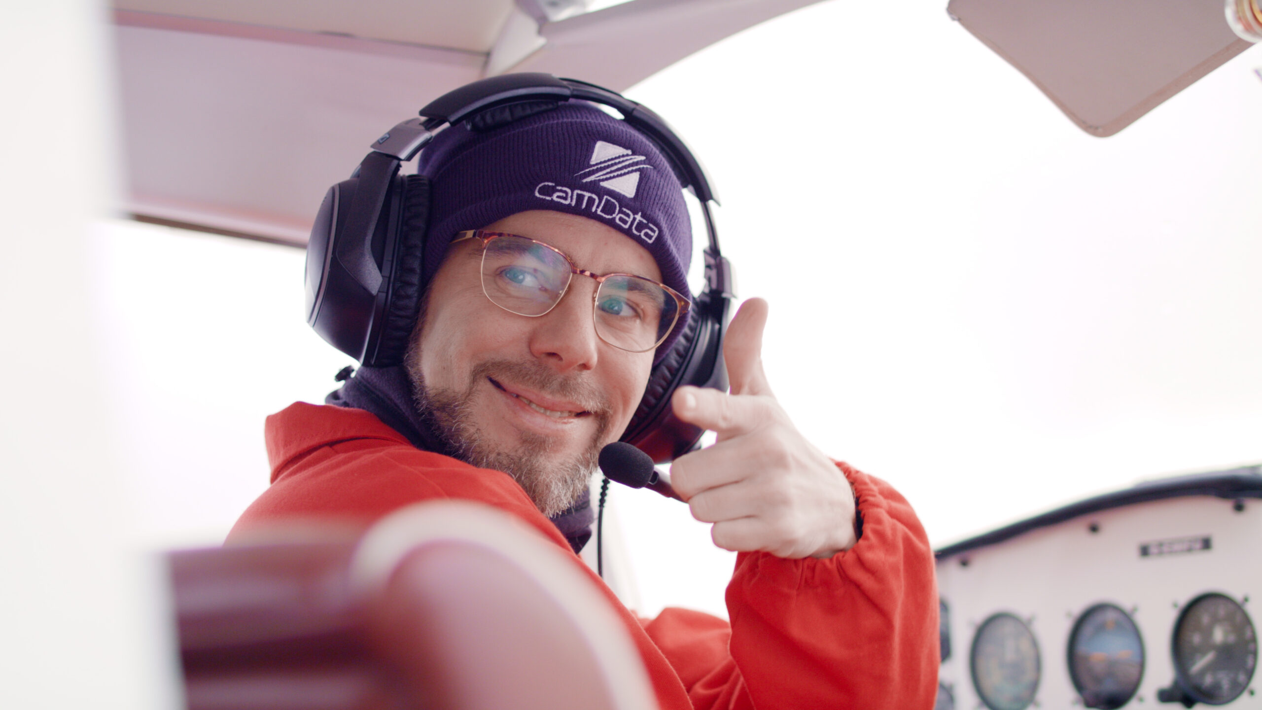 Mann mit Brille und Headset in Cockpit, lächelt und zeigt Fingerpistole, trägt Mütze und Jacke, Instrumente im Hintergrund.