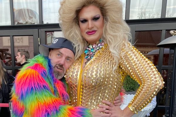 Zwei Personen posieren eng: eine in goldenem Outfit mit großer blonder Perücke, die andere in bunter Felljacke und Kappe.