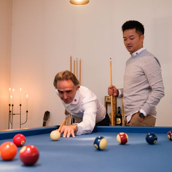Zwei Männer spielen Billard, einer stößt die Kugel an, der andere steht daneben und beobachtet.