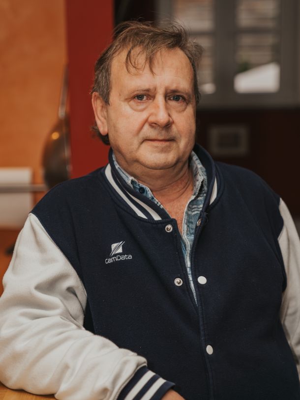 Mann in Jacke lehnt am Tisch, blickt ruhig in die Kamera, warm beleuchteter Innenraum mit entspannter Atmosphäre im Hintergrund.