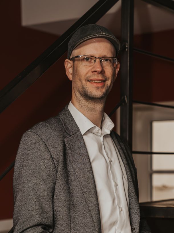 Mann mit Brille und Mütze im Sakko steht vor Treppe, lächelt in die Kamera, warmes Licht und moderner Innenraum im Hintergrund.